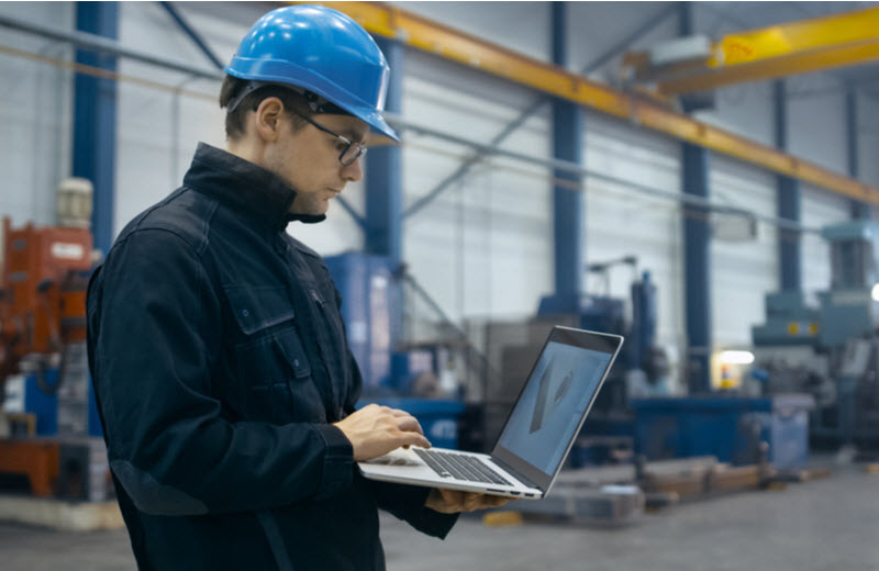 Factory worker in a hard hat is using a laptop computer with an engineering software