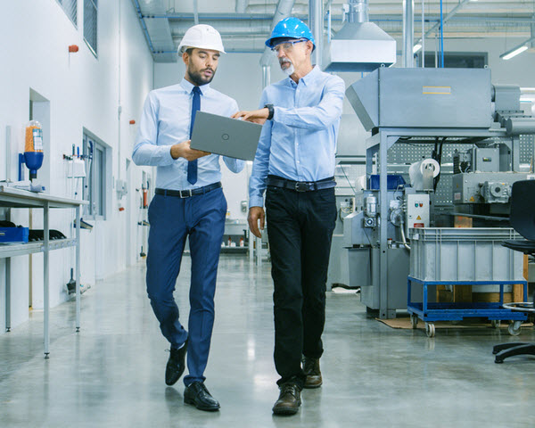 Head of the Project Holds Laptop and Discussing Product Details with Chief Engineer while They Walk Through Modern Factory